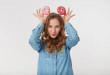 portrait of young cute male with long blond wavy hair wearing a denim shirt holds donuts like mickey mouse ears isoletad over white background