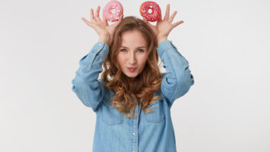 portrait of young cute male with long blond wavy hair wearing a denim shirt holds donuts like mickey mouse ears isoletad over white background