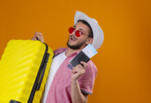 young handsome traveler guy wearing sunglasses in summer hat holding suitcase and air tickets looking confident and happy smiling cheerfully ready to travel standing over orange background
