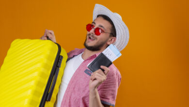 young handsome traveler guy wearing sunglasses in summer hat holding suitcase and air tickets looking confident and happy smiling cheerfully ready to travel standing over orange background