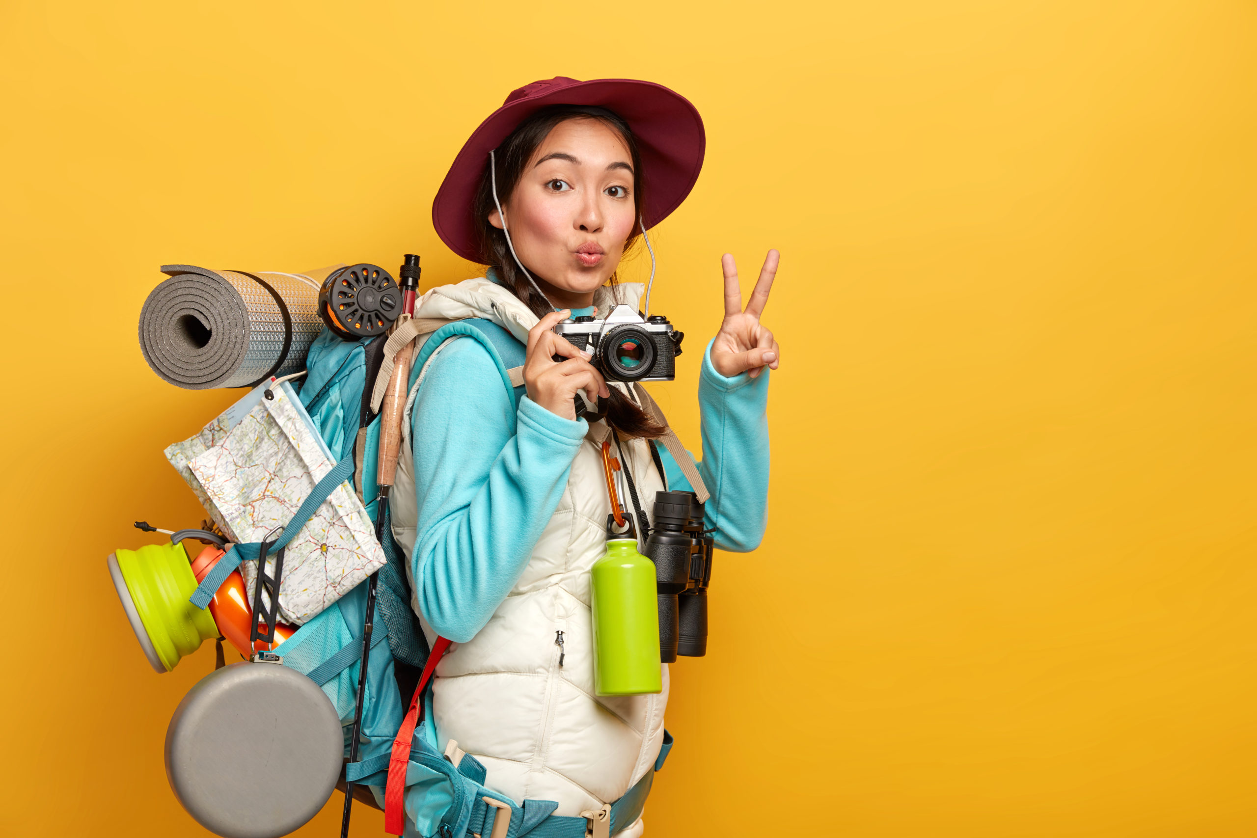 pretty active backpacker makes victory gesture keeps lips rounded holds retro camera stands with travelbag takes photos during trip isolated over yellow background scaled